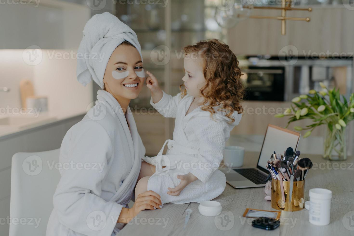 glad ung mamma lärare sin lilla dotter att göra makeup. culry liten flicka applicerar skönhetsplåster under ögonen på mamma använder skönhetsprodukter naturl kosmetika. hudvård och familjetid koncept foto