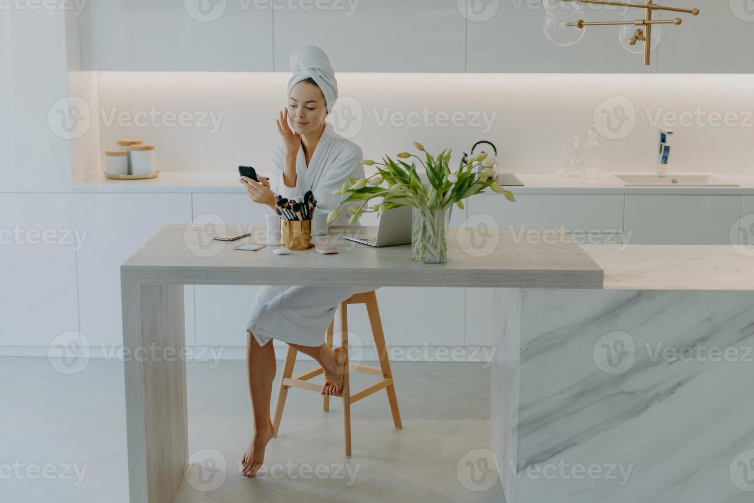vacker ung kvinnlig modell poserar i modern lägenhet bär badrock och handduk på huvudet applicerar ansiktskräm genomgår skönhetsprocedurer använder kosmetiska verktyg för hudvård. välbefinnande foto