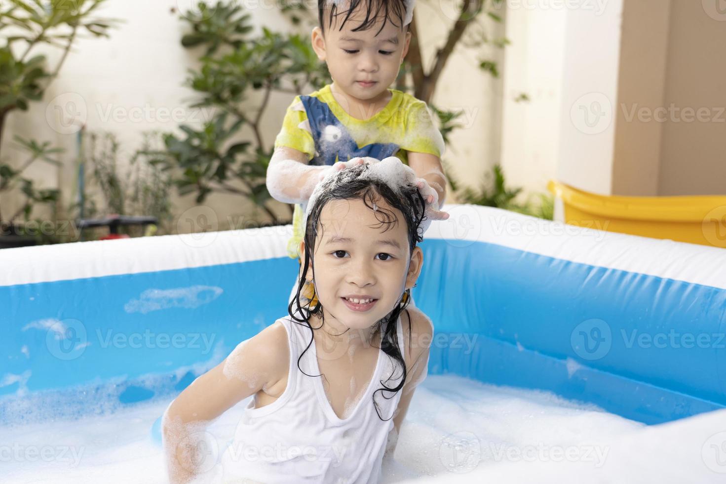 de liten bror var tvättning hans systerns hår i ett uppblåsbar slå samman. flicka och pojke spelar med tvål bubblor i de simning slå samman på Hem. Lycklig till spela i de vatten under de sommar semester. foto
