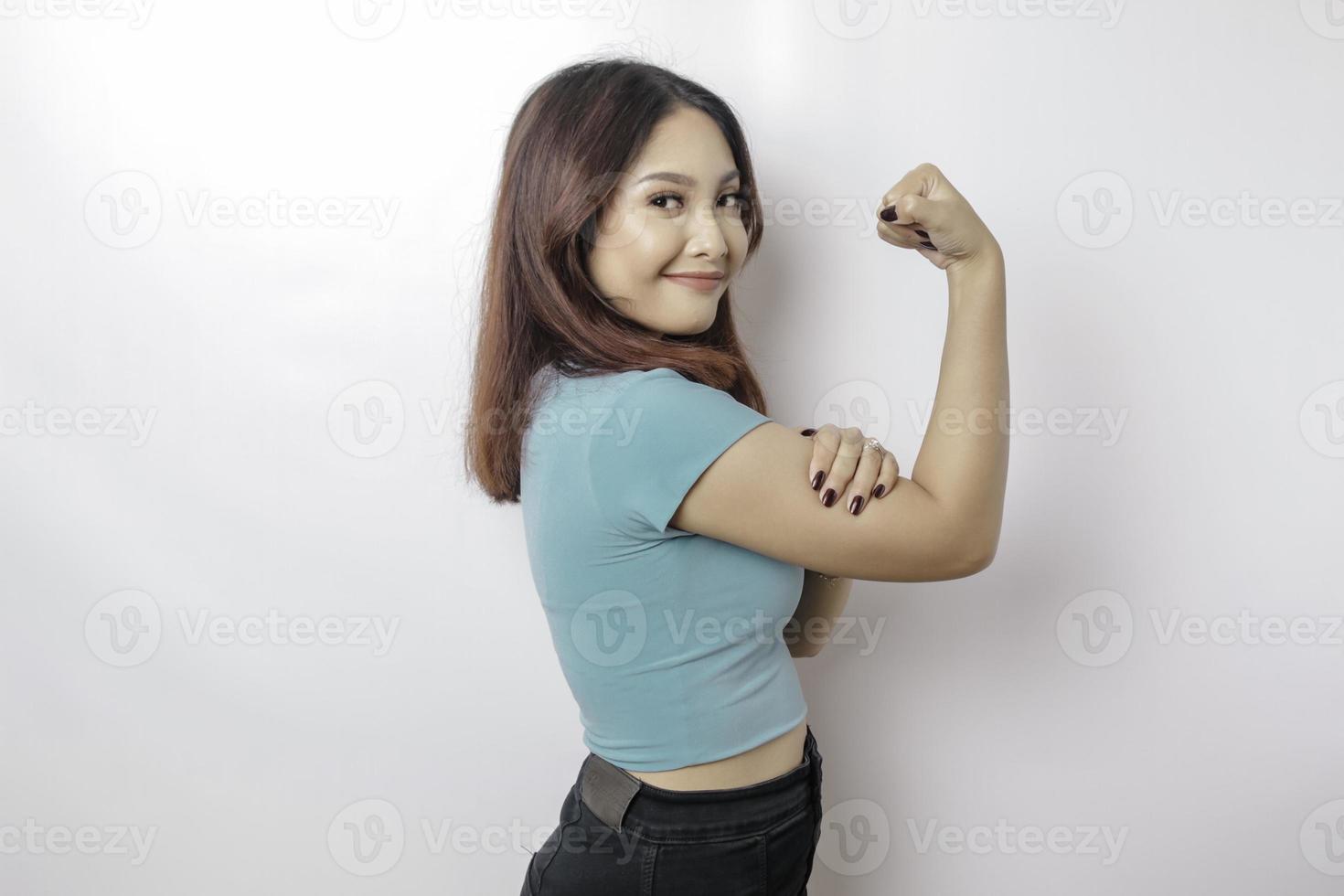 upphetsad asiatisk kvinna bär en blå t-shirt som visar stark gest förbi lyft henne vapen och muskler leende stolt foto