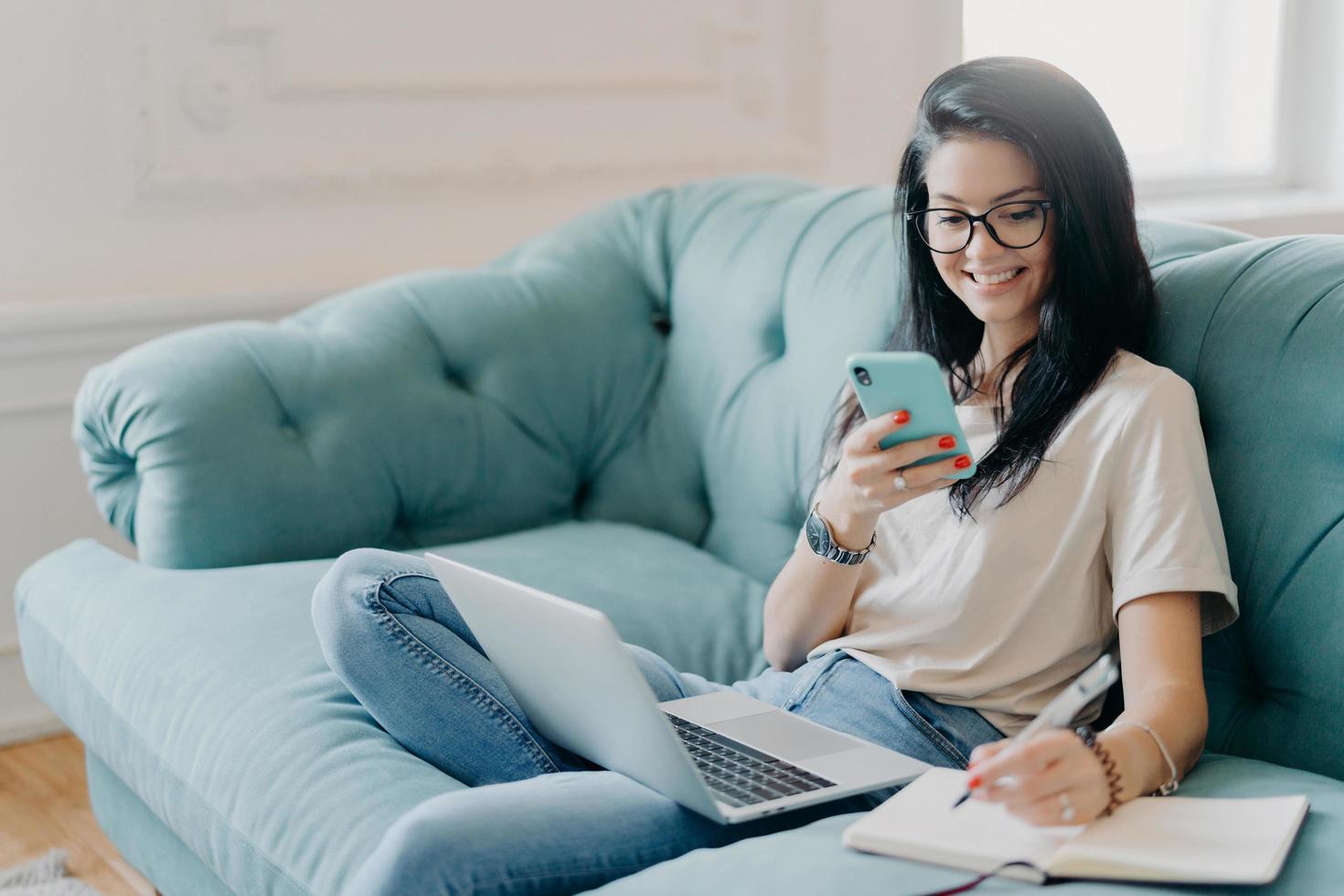 skicklig kvinnlig student gör presentation för projekt, söker nödvändig information via mobiltelefon, har öppnat laptop på knä, bär vardagskläder, poserar på soffan i mysiga rum, chattar online foto