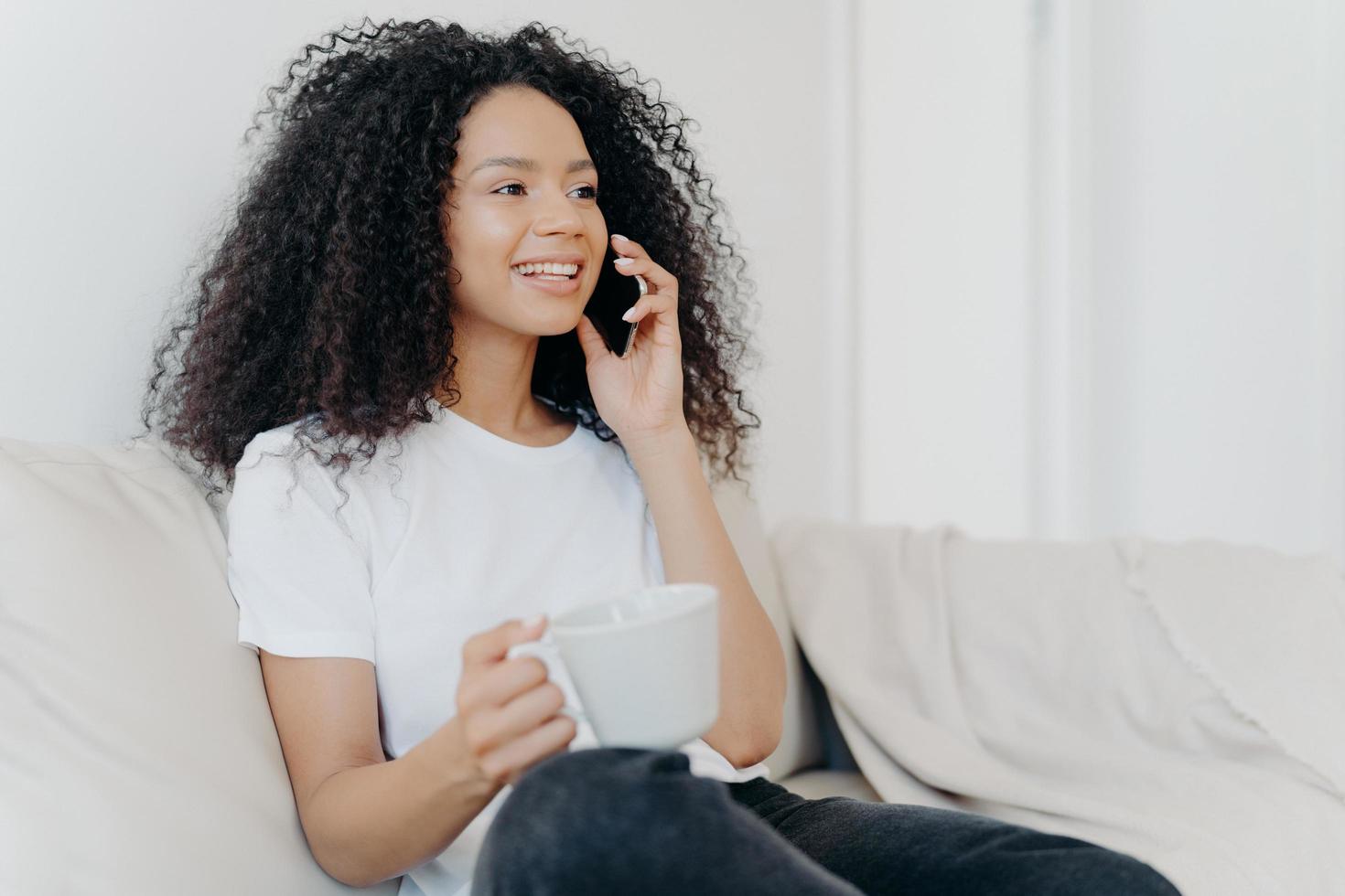 skott i sidled av positiv etnisk brunett kvinna har telefonsamtal, dricker varm dryck, håller en vit kopp, sitter på bekväm soffa i vardagsrummet, hör trevliga nyheter, ler glatt foto