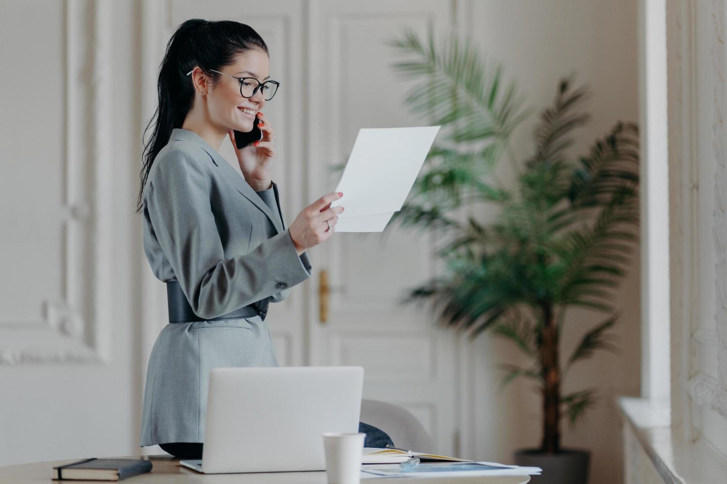 inomhusbild av glad leende kvinna chef kontrollerar information från tidningar bär grå formell kostym, optiska glasögon koncentrerade på forskningsarbete har telefonsamtal, står i coworking space foto