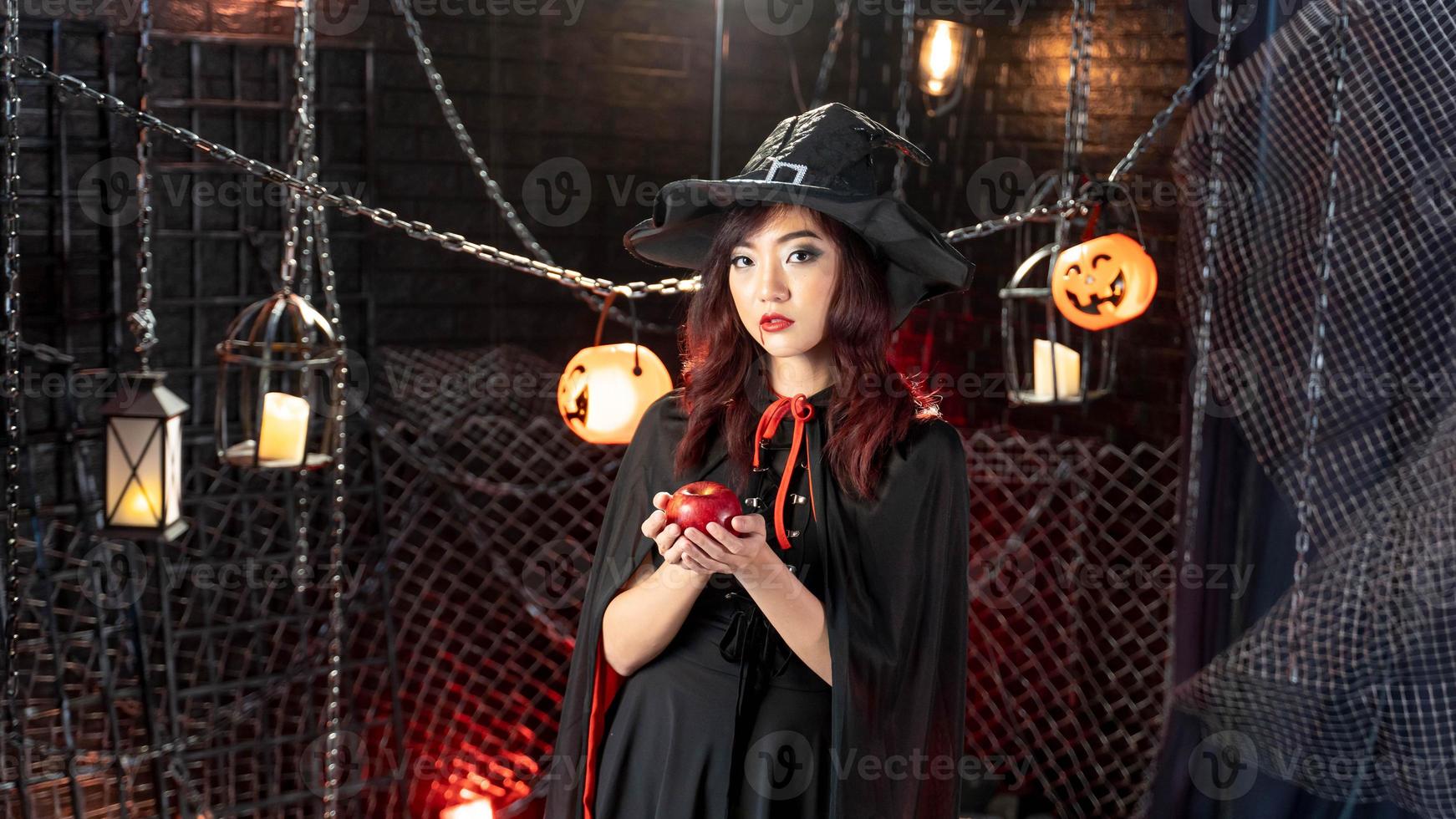 Lycklig halloween, ung asiatisk kvinna i häxa kostym bär häxa hatt innehav äpple i halloween tema. foto