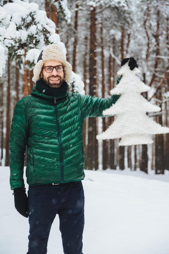 porträtt av glad skäggig stilig man bär varm hatt, grön anorak och handskar håller liten vit konstgjord gran, står i vinterskog täckt med snö. vackra landskap foto