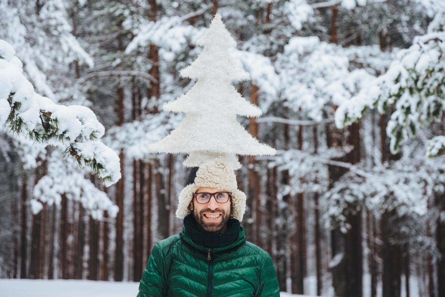 horisontell bild av leende manlig modell håller gran på huvudet, står utanför i vinterskogen, njuter av lugn och lugn atmosfär. kaukasisk orakad man med glada uttryck foto