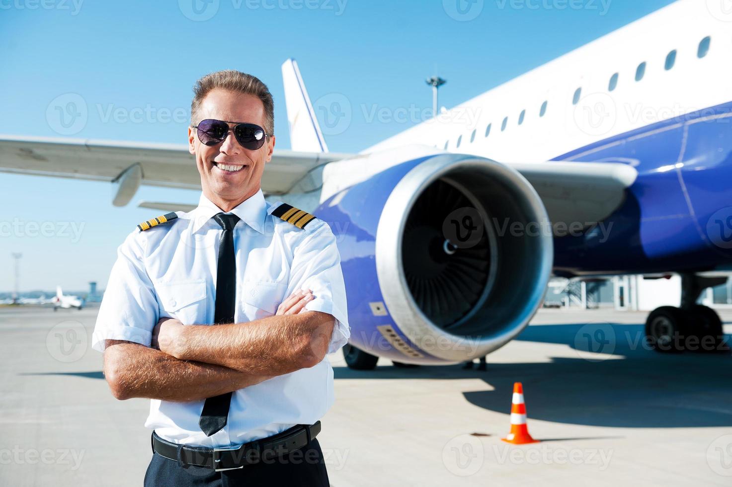 självsäker pilot. självsäker manlig pilot i enhetlig förvaring vapen korsade och leende med flygplan i de bakgrund foto