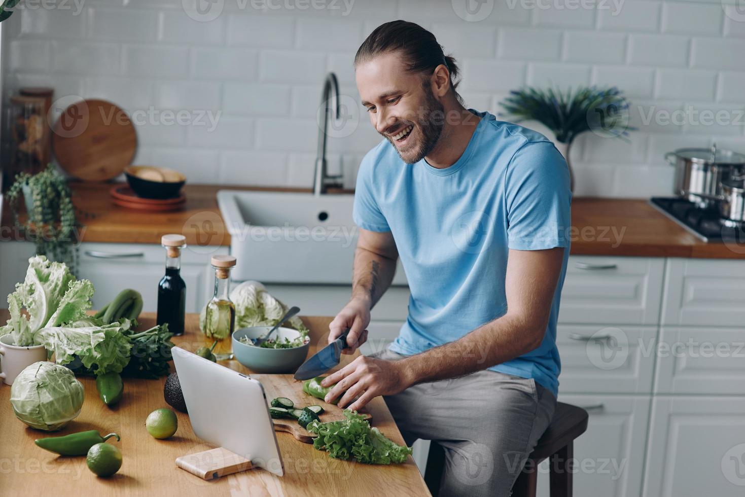Lycklig ung man ser på digital läsplatta medan framställning mat på de inhemsk kök foto