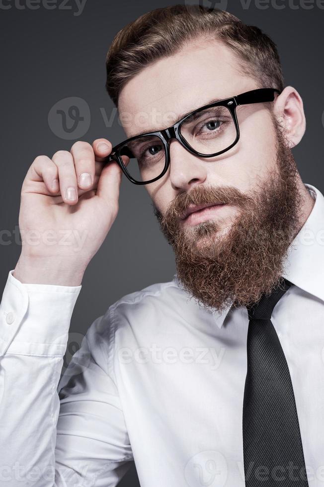 han fick kreativ sinne. stilig ung skäggig man i skjorta och slips justeras hans glasögon och ser på kamera medan stående mot grå bakgrund foto