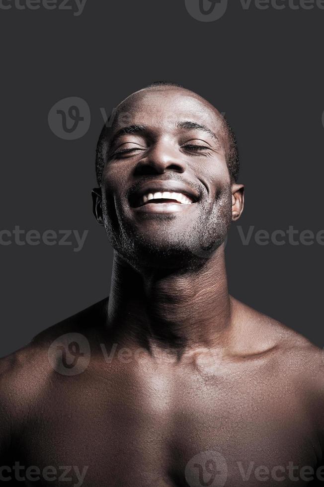 uppriktig lycka. porträtt av ung bar överkropp afrikansk man förvaring ögon stängd och leende medan stående mot grå bakgrund foto