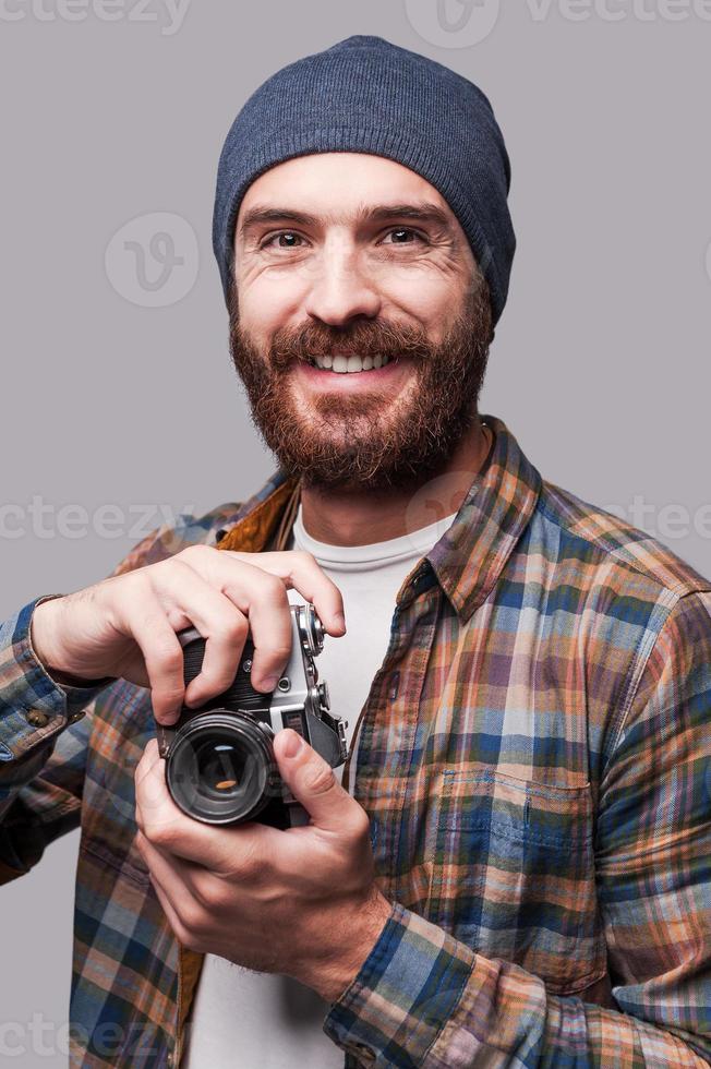 självsäker fotograf. stilig ung skäggig man innehav gammaldags kamera och leende medan stående mot grå bakgrund foto