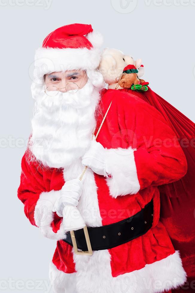 glad jul traditionell santa claus bärande säck med presenterar och ser på kamera medan stående mot grå bakgrund foto