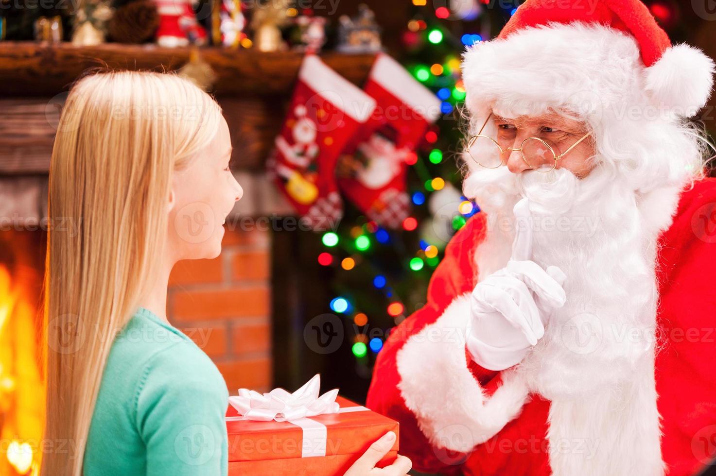 du gjorde inte ser mig. Lycklig liten flicka innehav gåva låda och ser på verklig santa claus gestikulerar tystnad tecken med jul träd och öppen spis i de bakgrund foto