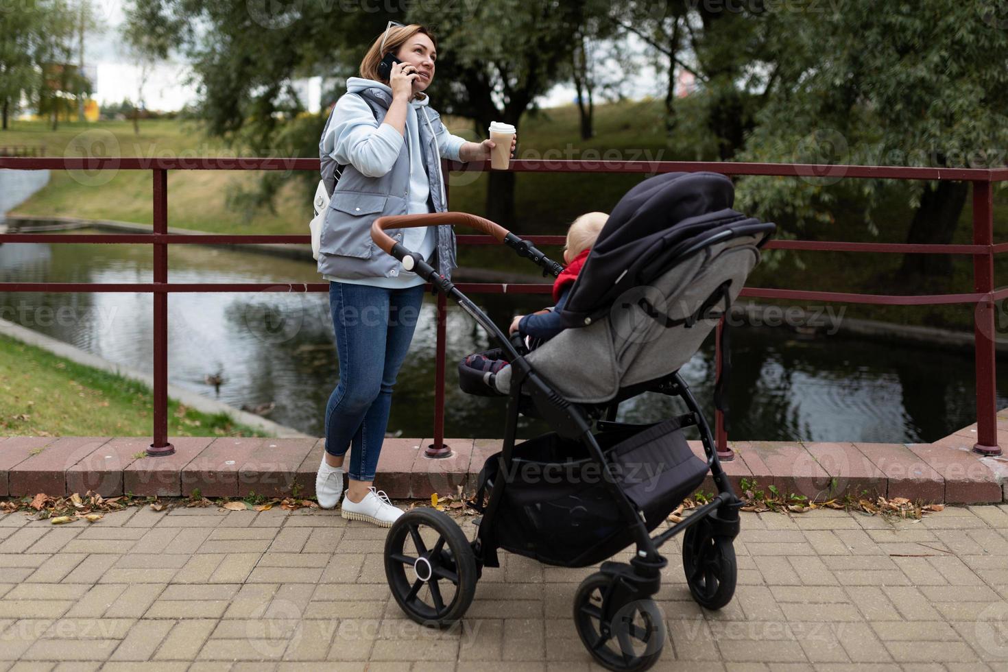 ung omtänksam mor med en kopp av kaffe i henne händer Nästa till de sittvagn med henne nyfödd barn medan gående i de parkera talar på de telefon med en leende på henne ansikte foto