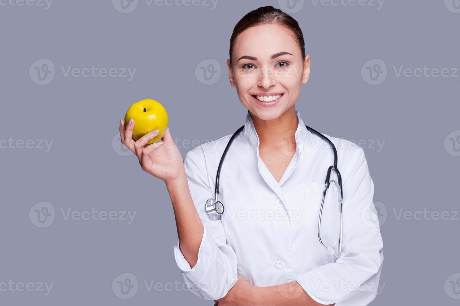äta friska självsäker kvinna läkare i vit enhetlig innehav grön äpple och leende medan stående mot grå bakgrund foto