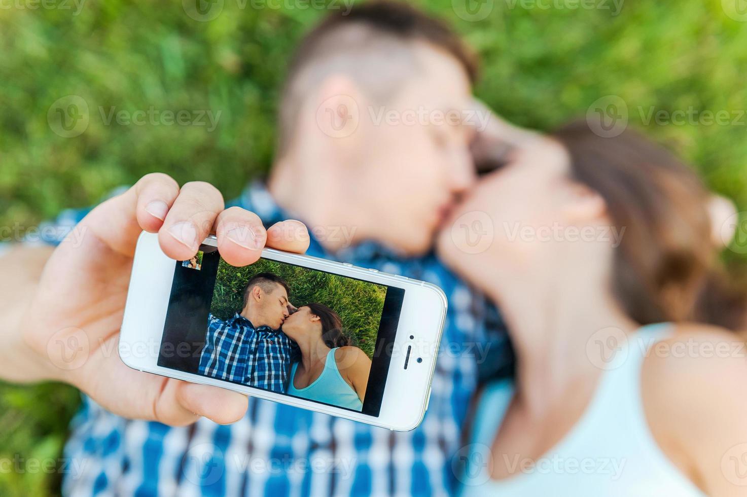 fångande en ögonblick. topp se av Lycklig ung kärleksfull par framställning selfie med smart telefon medan liggande på de gräs och kissing foto