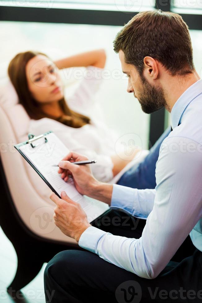 psykiater på arbete. självsäker manlig psykiater skrivning något på hans Urklipp medan kvinna Sammanträde i de bakgrund foto