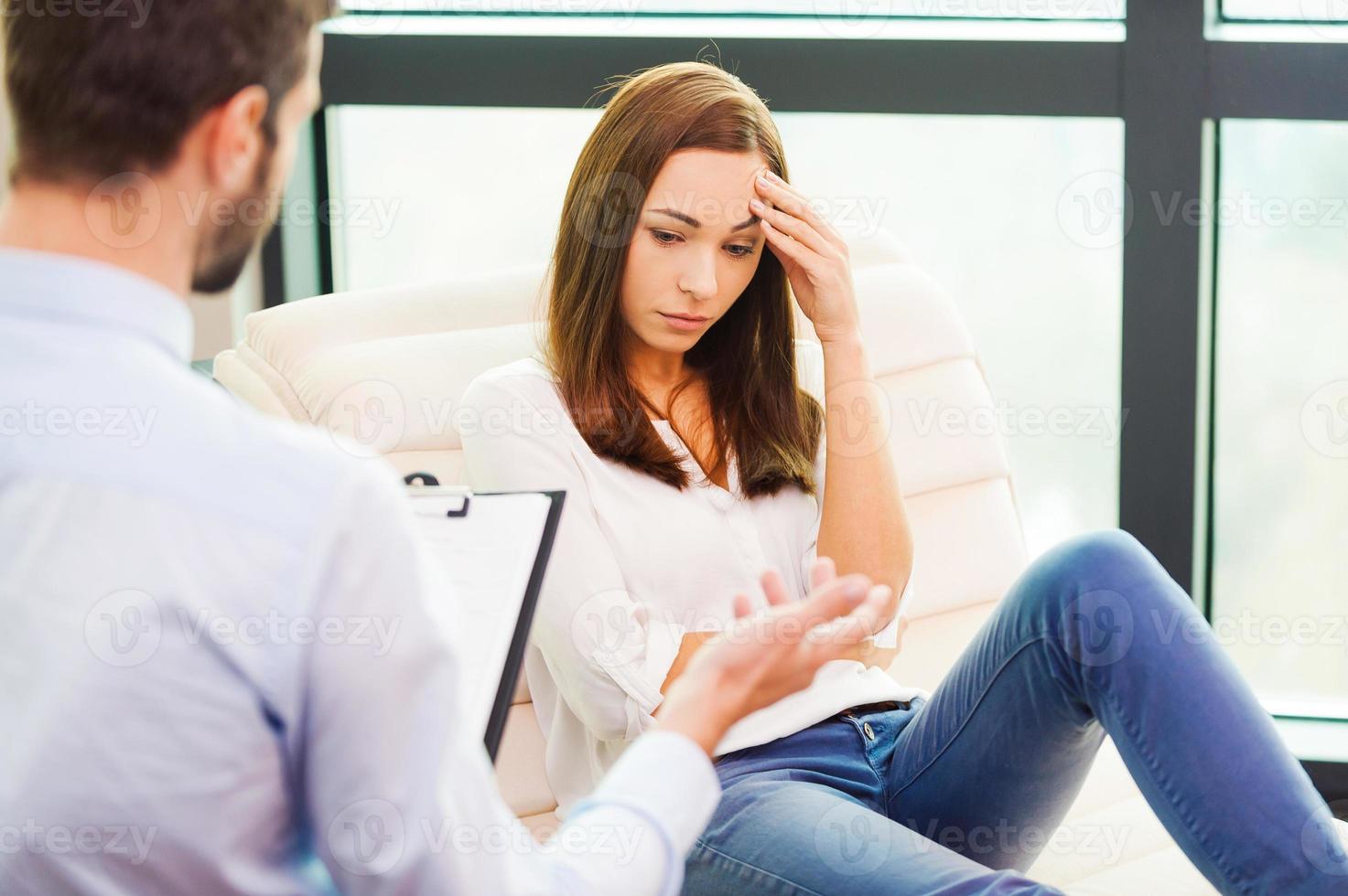 känsla så hopplös. deprimerad ung kvinna Sammanträde på de stol och innehav hand på huvud medan manlig psykiater Sammanträde i främre av henne och innehav Urklipp foto