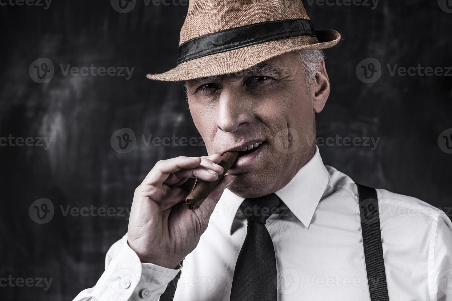 stor chef. allvarlig senior man i hatt och hängslen rökning cigarr och ser på du medan stående mot mörk bakgrund foto