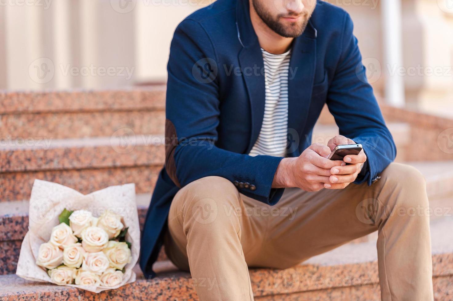 skriver meddelande till flickvän. närbild av stilig ung man i smart jacka ser på de mobil telefon och medan Sammanträde på de trappa med bukett av ro om nära honom foto