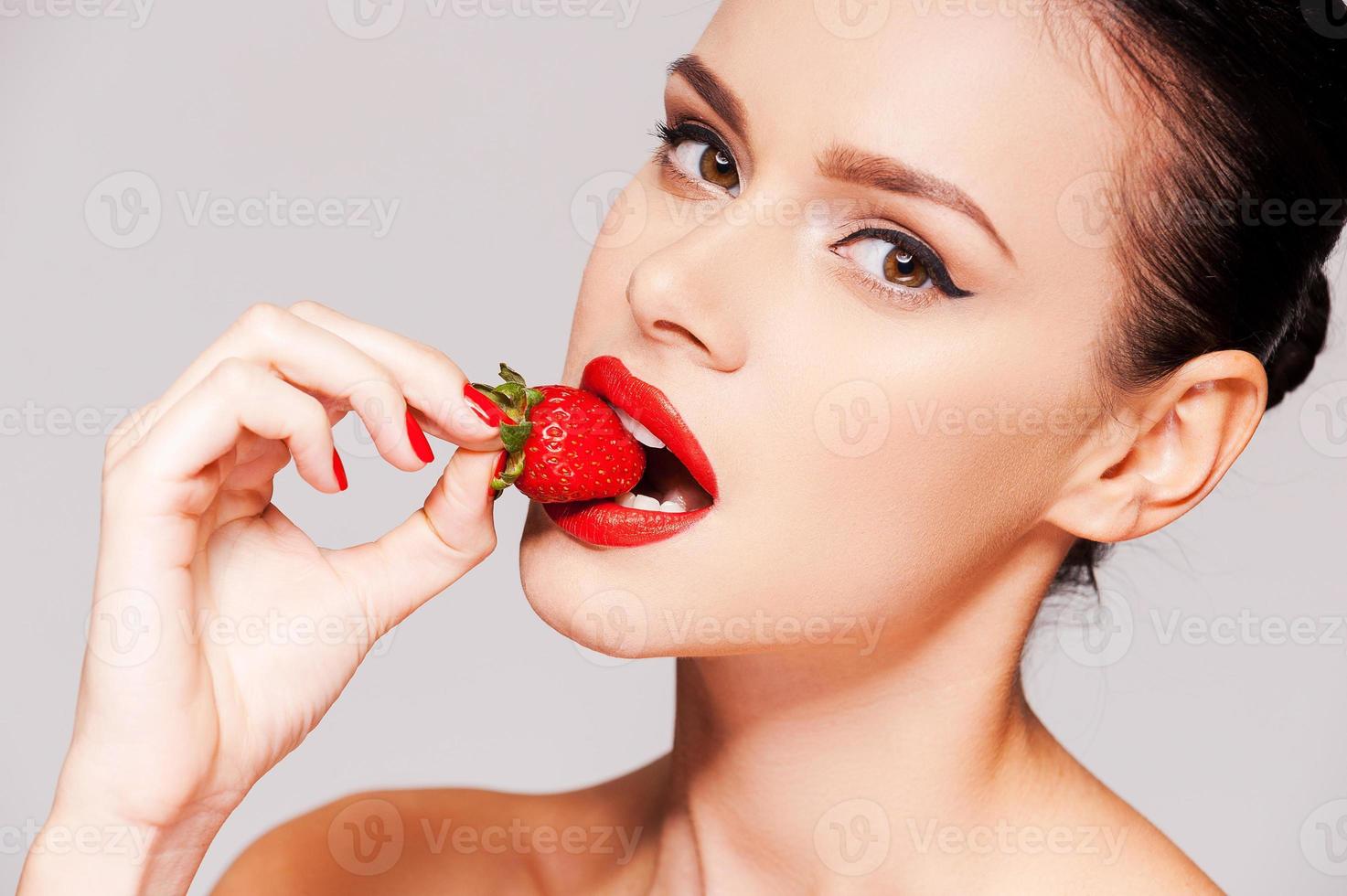 jordgubb mun. skön ung bar överkropp kvinna innehav jordgubb i henne hand och provsmakning den medan stående mot grå bakgrund foto