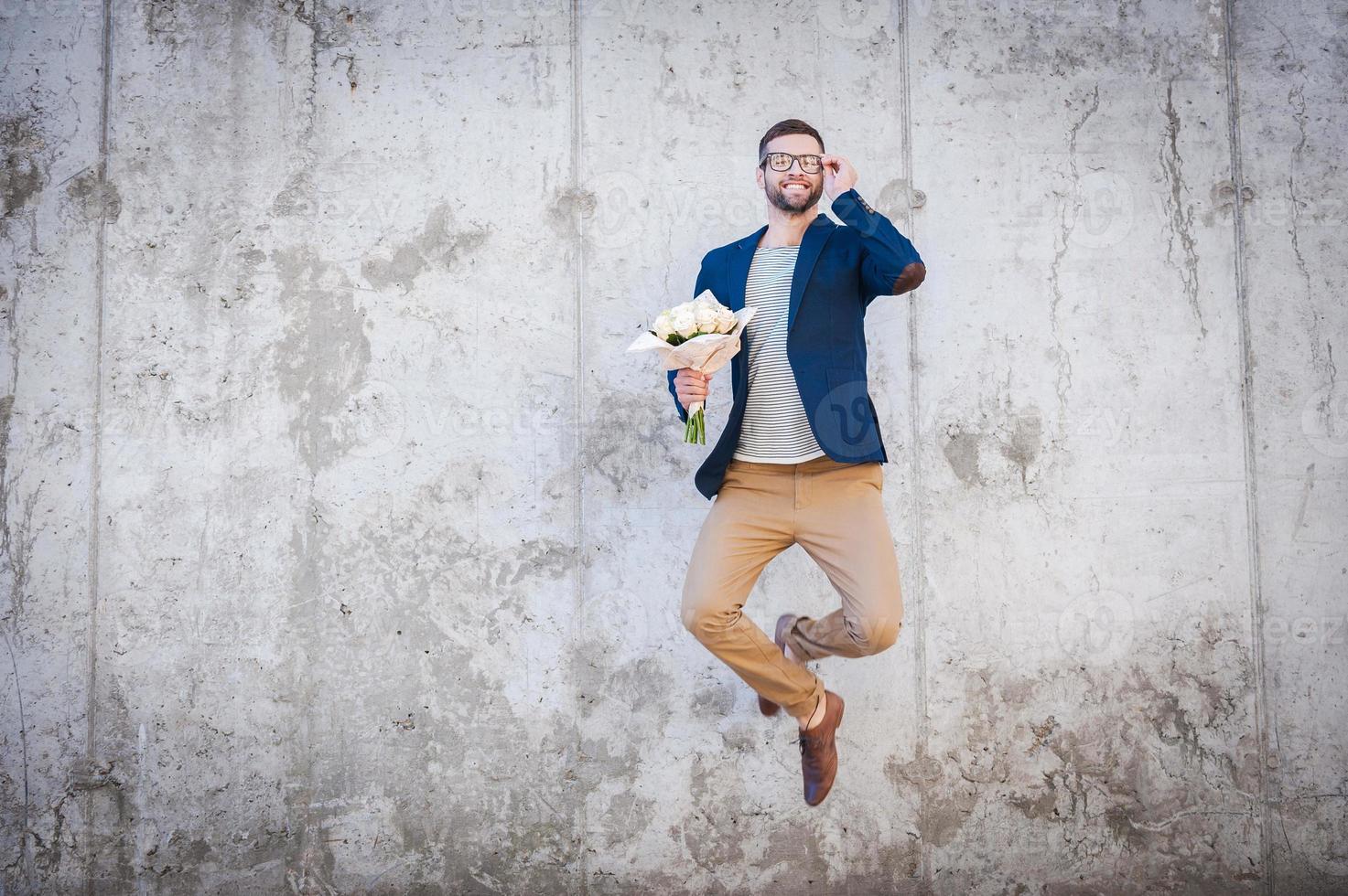 Lycklig och i kärlek. Lycklig ung man i smart jacka innehav bukett av blommor och Hoppar i främre av de betong vägg foto