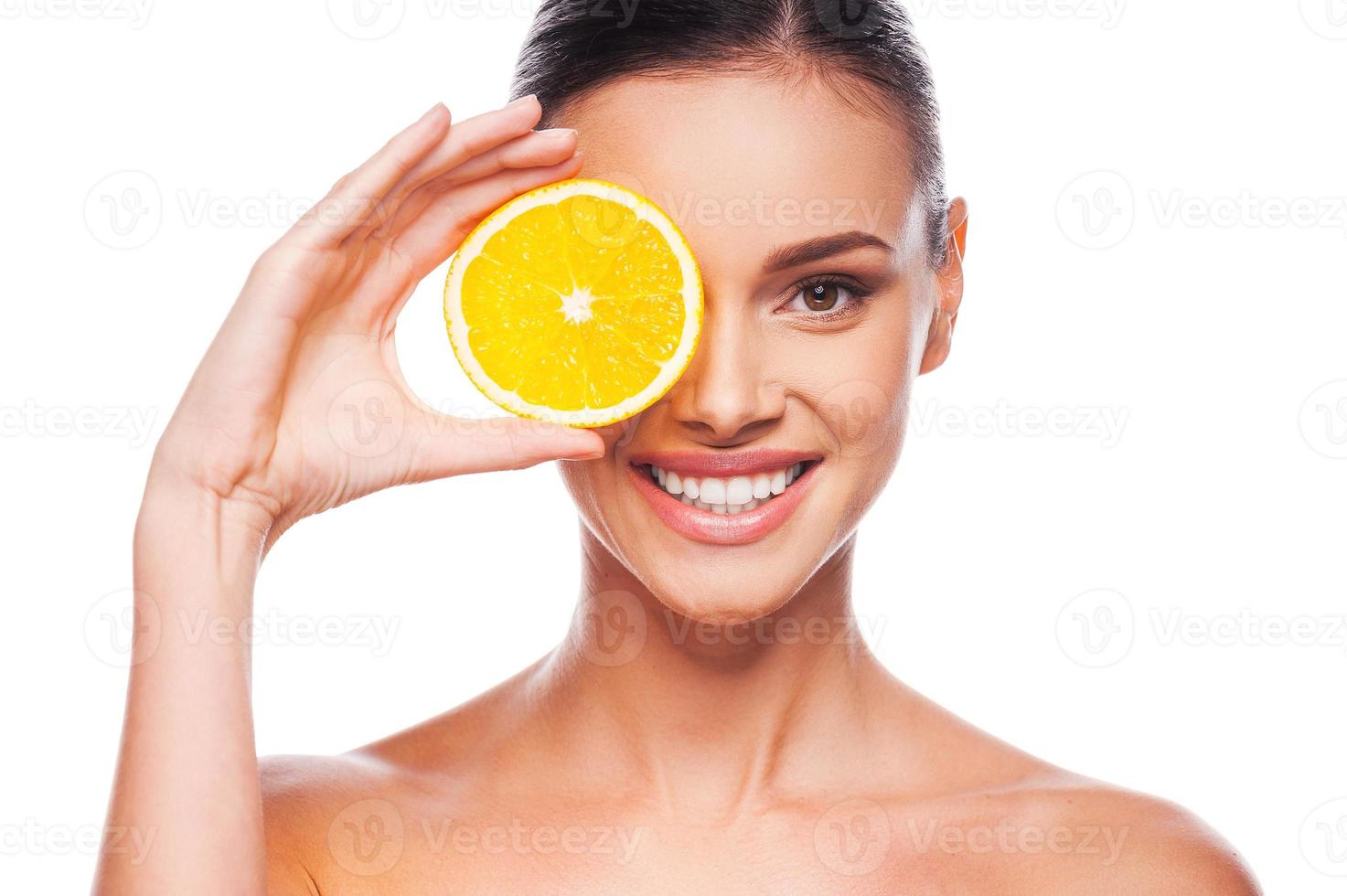 bra mat för en friska livsstil. skön ung bar överkropp kvinna innehav bit av orange i främre av henne öga medan stående mot vit bakgrund foto