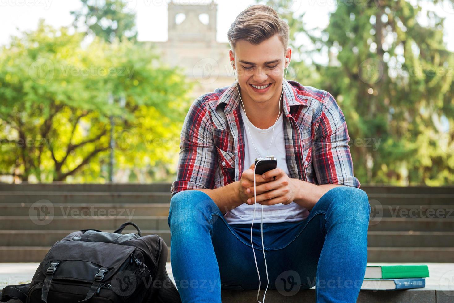 njuter hans favorit musik. Lycklig manlig studerande lyssnande till mp3 spelare och leende medan Sammanträde på de utomhus trappa med böcker och ryggsäck om nära honom foto