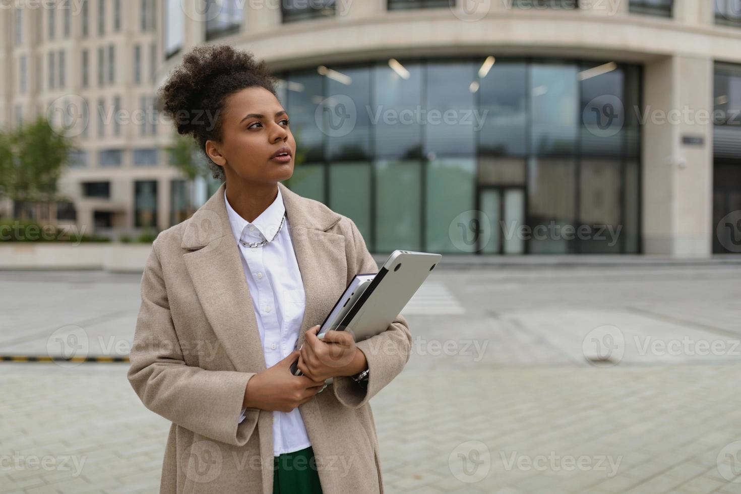porträtt av en ung företag afrikansk amerikan kvinna med en bärbar dator i henne händer mot de bakgrund av en icke-moderna administrativ stad byggnad foto