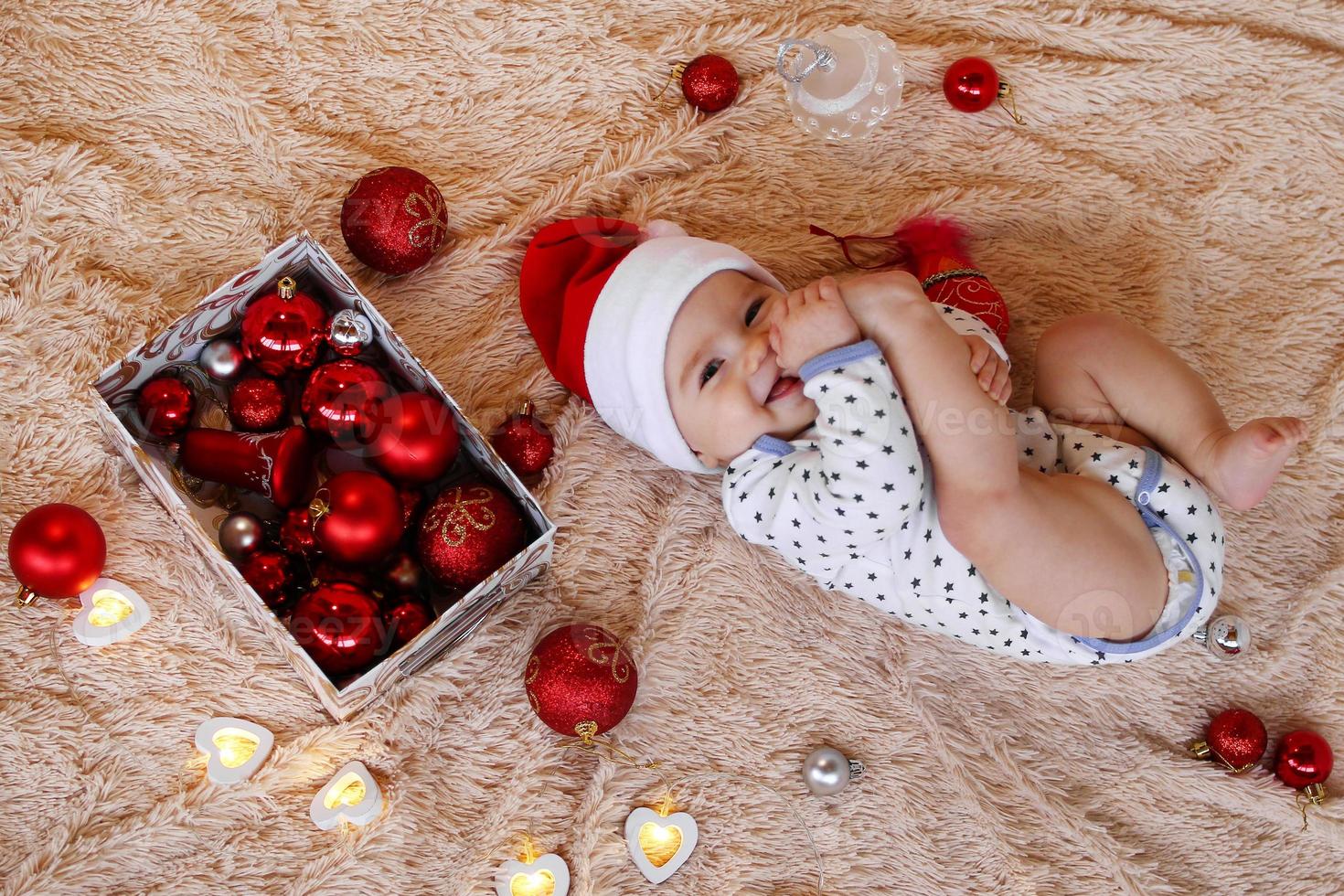 leende liten flicka i santa claus röd hatt är om på en beige pläd med röd och vit jul dekorationer och jul lampor och innehav henne fot i henne mun, topp se. foto