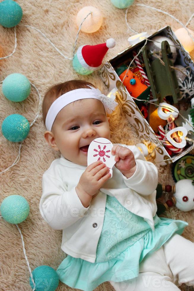 söt liten flicka i blå och vit klänning är spelar med trä- leksak på en beige pläd med jul dekorationer och lampor, topp se. foto