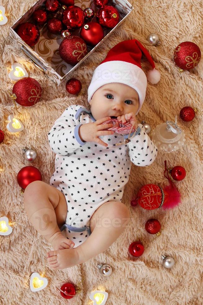 förtjusande liten flicka i santa claus röd hatt är spelar med trä- leksak på en beige pläd med röd och vit jul dekorationer och jul lampor, topp se. foto