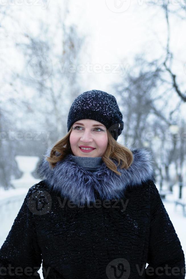 porträtt av ung skön kvinna med kort blond hår i svart falsk päls täcka med grå fluffig huva och svart stickat hatt är gående i vinter- snöig parkera. foto