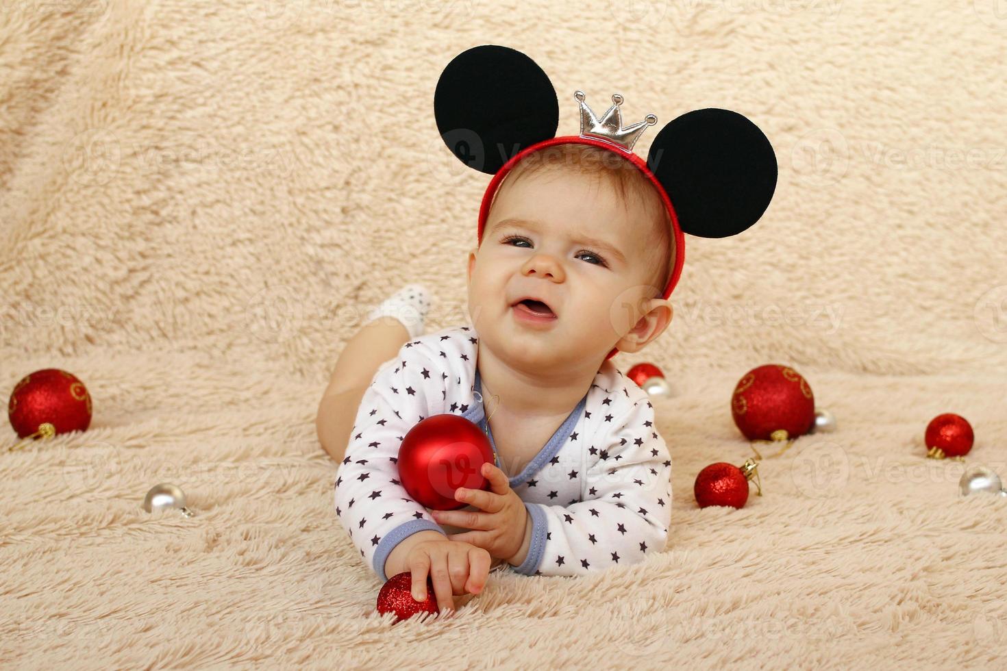 porträtt av söt liten flicka med mus öron på en beige pläd och röd skinande jul bollar. foto