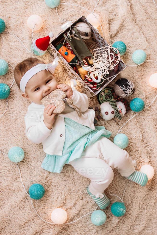 skön leende liten flicka i blå och vit klänning är spelar med trä- hjärta på en beige pläd med jul dekorationer och lampor, topp se. foto
