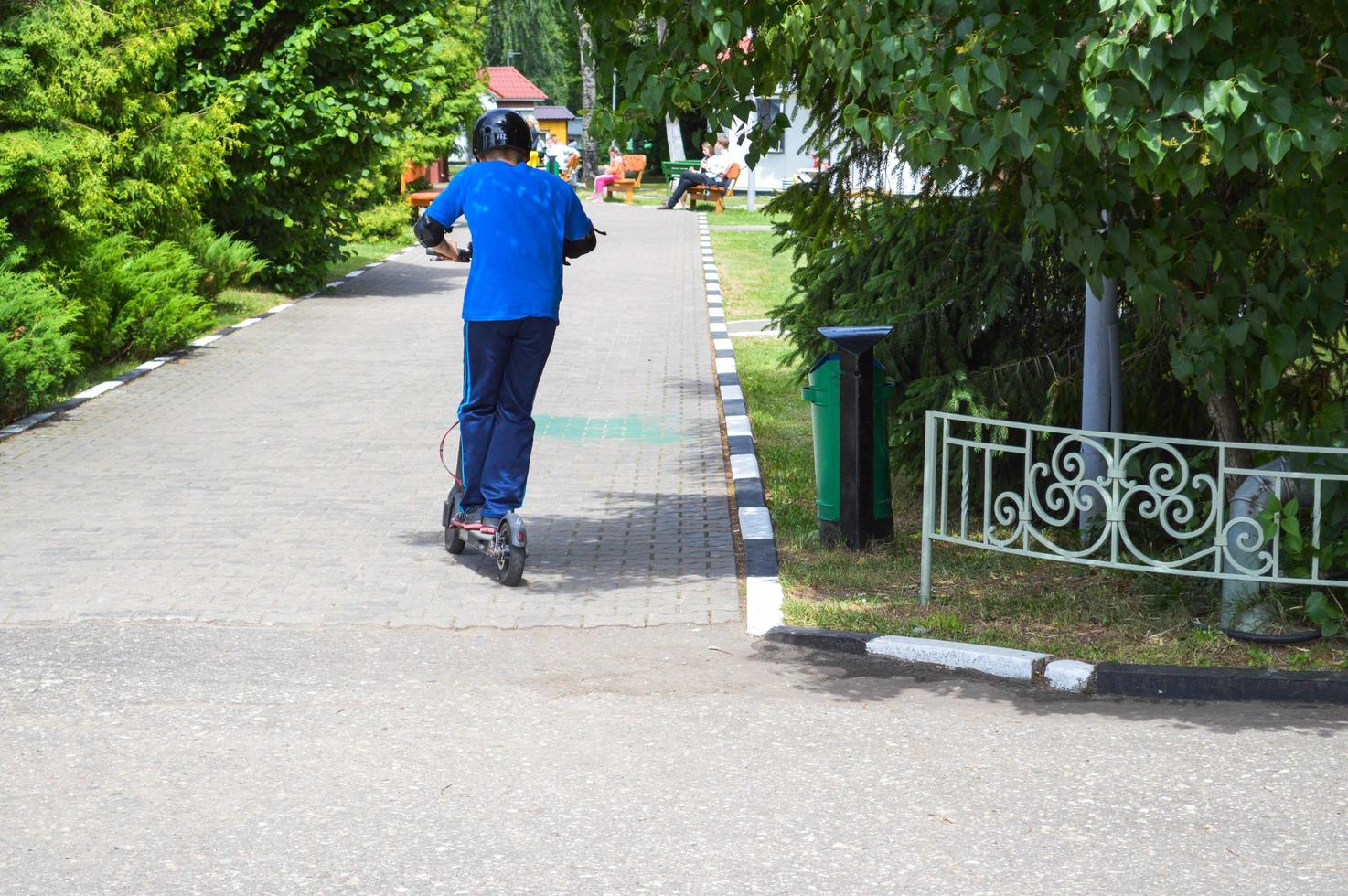 en pojke, en man rider längs de parkera väg på en ny modern elektrisk skoter på batterier, stående på den med två ben foto