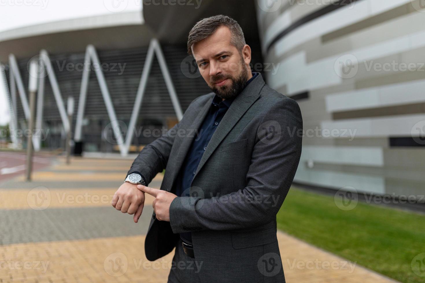 ett vuxen man i en företag kostym poäng hans finger på de klocka på de bakgrund av ett kontor byggnad foto