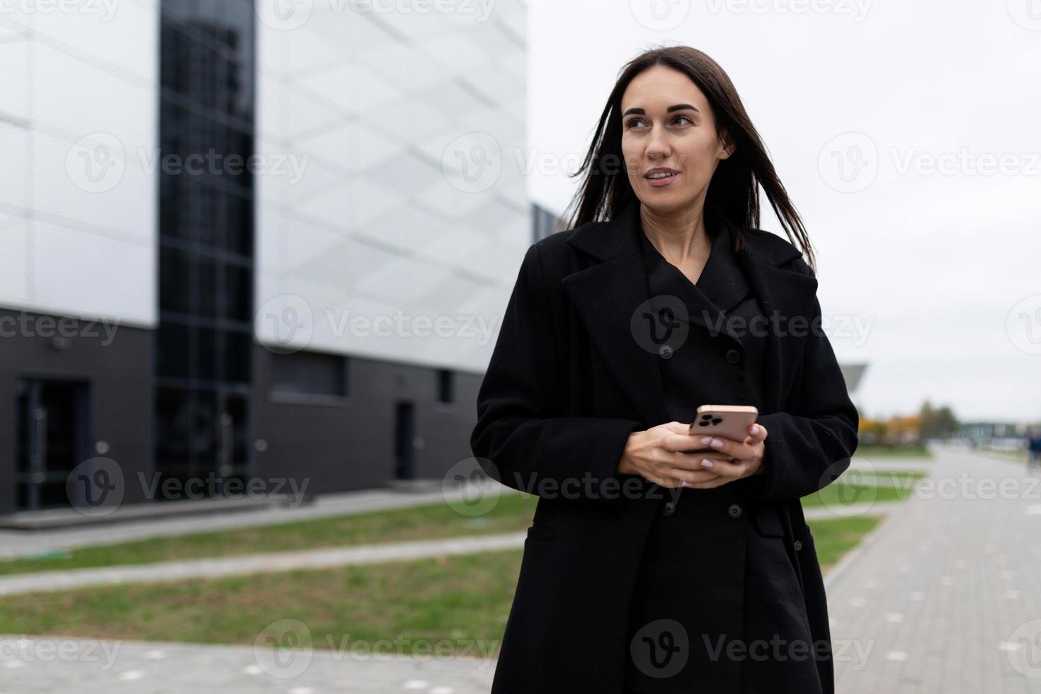 företag eleganta kvinna i en svart täcka med en mobil telefon på de bakgrund av en modern byggnad foto