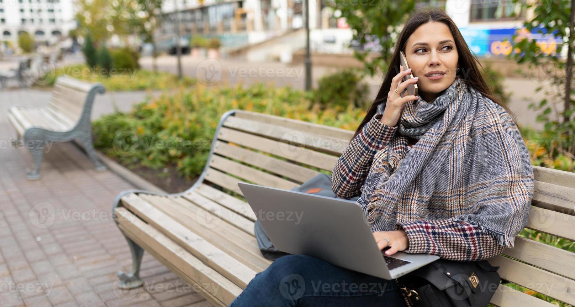kvinna frilansare arbetssätt uppkopplad på bärbar dator medan Sammanträde i stad på trä- bänkar foto