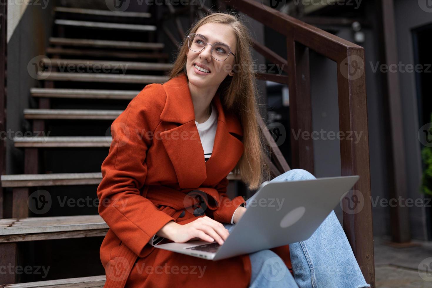 ung kvinna arbetssätt på en bärbar dator uppkopplad Sammanträde på en flyg av trappa Nästa till en Kafé leende ser bort foto