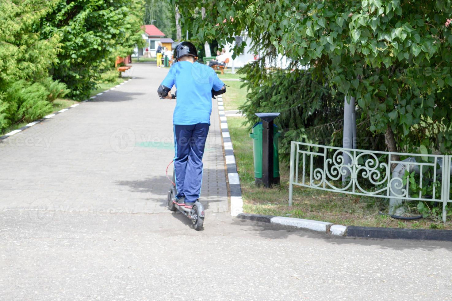 en pojke, en man rider längs de parkera väg på en ny modern elektrisk skoter på batterier, stående på den med två ben foto