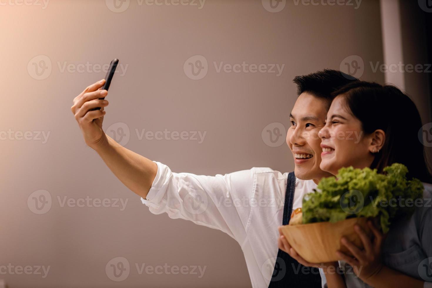 en leende par av manlig och kvinna använder sig av telefon i de kök. tar selfie eller leva strömning via smartphone medan framställning en vegan mat. modern friska livsstil begrepp. matlagning på Hem tillsammans foto