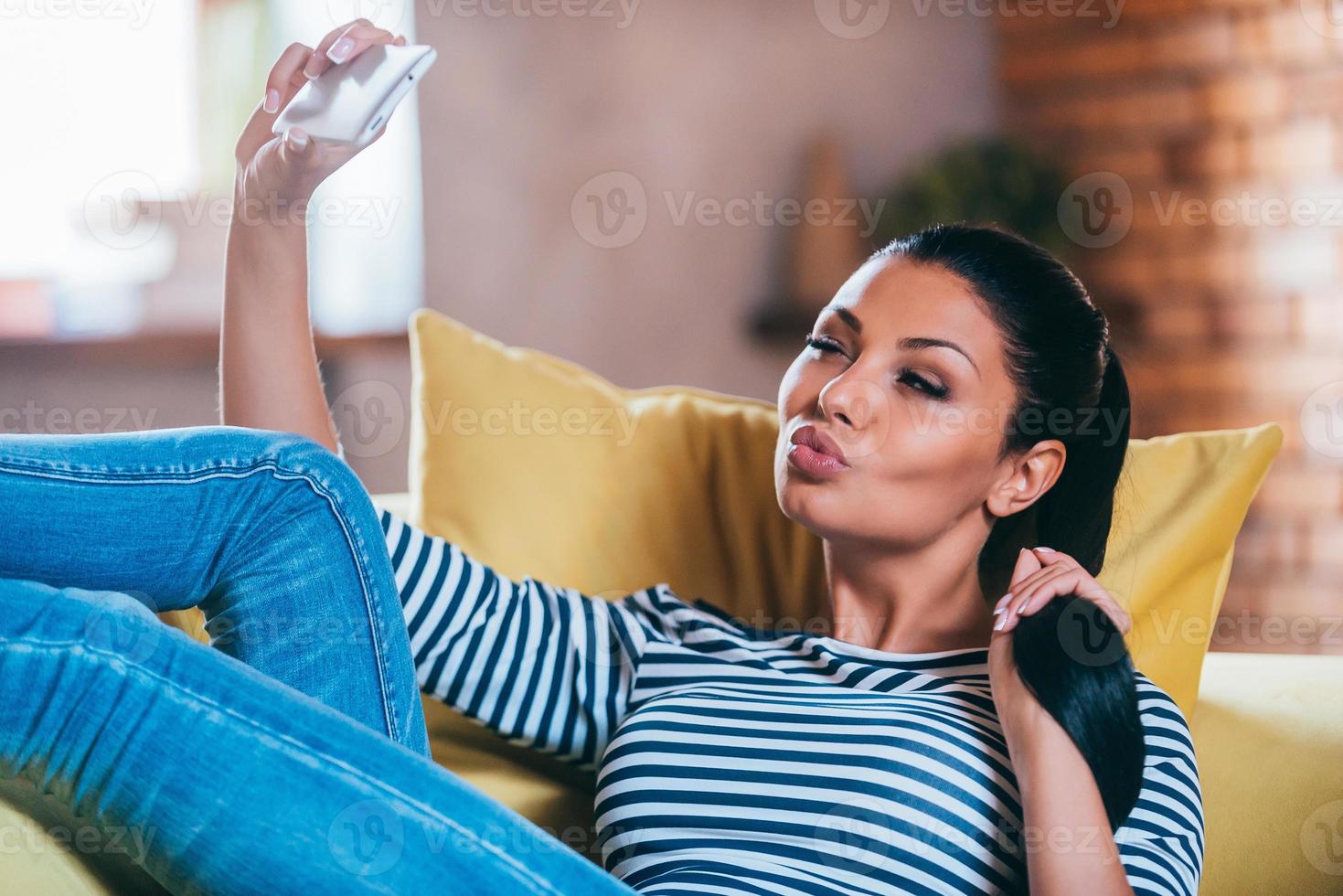 selfie tid. skön ung kvinna framställning selfie förbi henne smart telefon och framställning en ansikte medan Sammanträde på de soffa på Hem foto