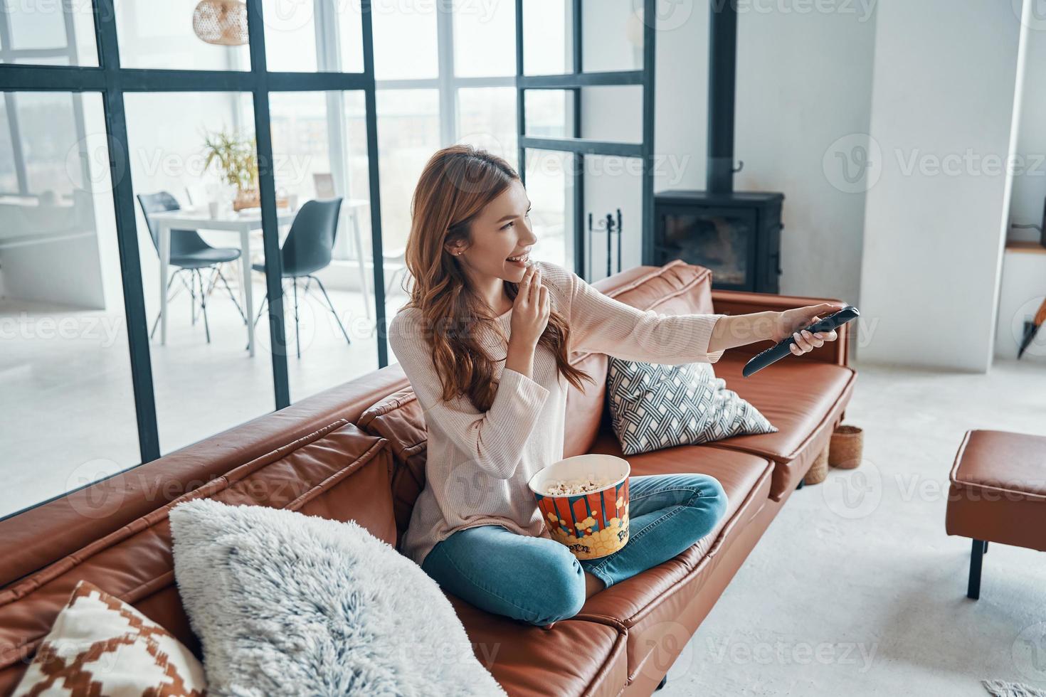 skön ung kvinna tittar på tv och äter popcorn medan Sammanträde på de soffa på Hem foto