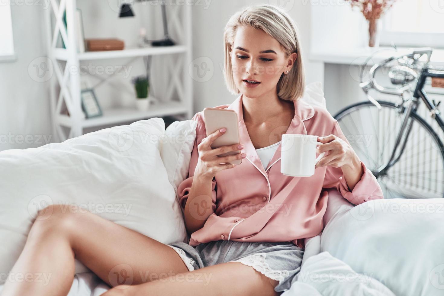 chattar med vänner. attraktiv ung kvinna använder sig av smart telefon medan Sammanträde på säng på Hem foto