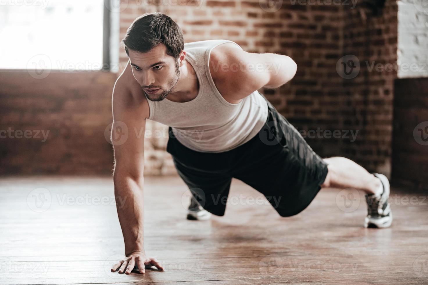 ett hand tryck upp. självsäker muskulös ung man bär sport ha på sig och håller på med ett hand tryck upp medan utövar på de golv i loft interiör foto