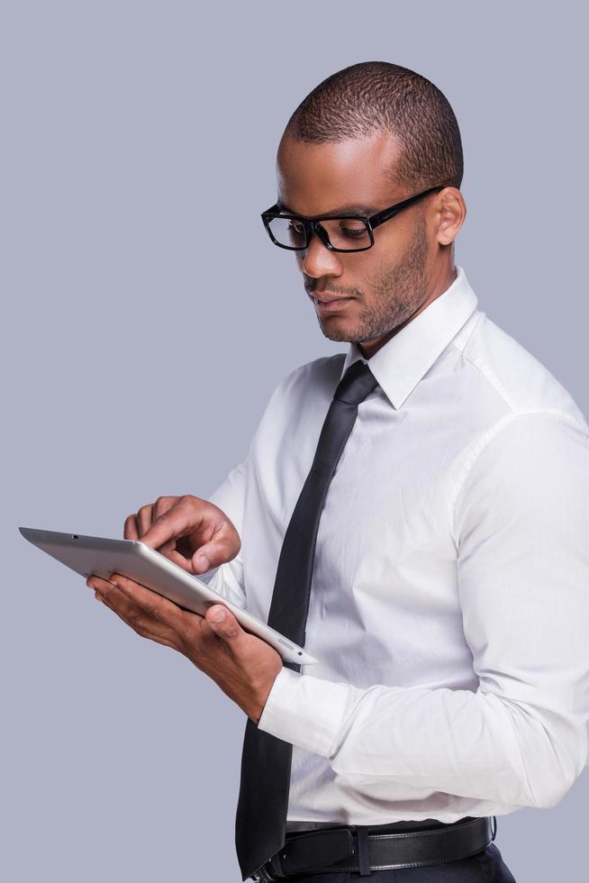 granskning hans ny grej. självsäker ung afrikansk man i skjorta och slips arbetssätt på digital läsplatta medan stående mot grå bakgrund foto