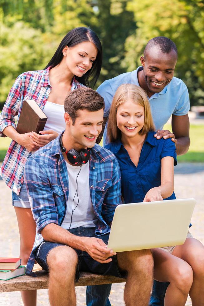 surfing webb tillsammans. fyra Lycklig ung människor diskuterar något och ser på de bärbar dator medan Sammanträde utomhus tillsammans foto