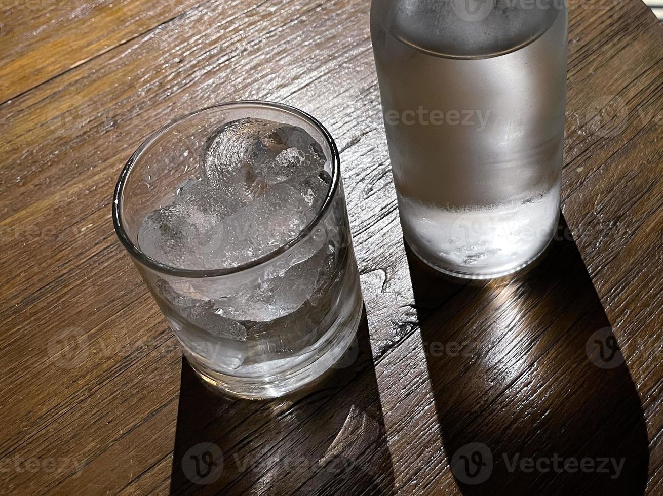 selektiv fokus, en klar glas fylld med is och klar glas flaska innehar kall mineral vatten sitter på trä- tabell foto