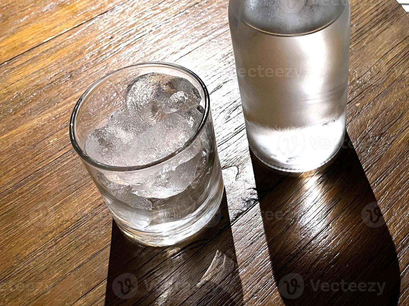 selektiv fokus, en klar glas fylld med is och klar glas flaska innehar kall mineral vatten sitter på trä- tabell foto